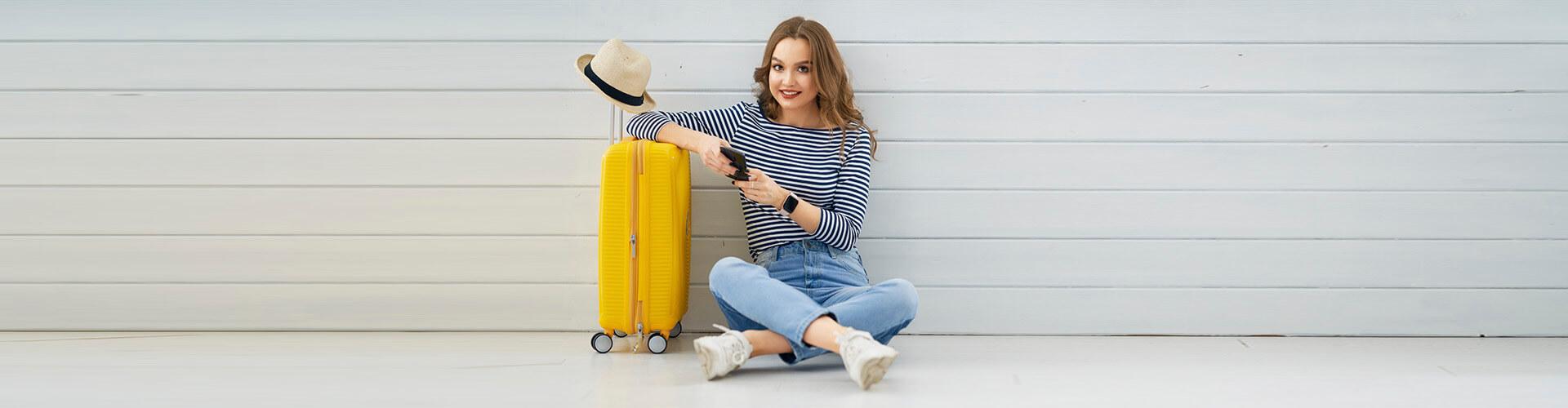 Segur d'Assistència en Viatge - Dona jove asseguda en el sòl amb el mòbil a la mà, al costat de la seua maleta i un barret preparant un viatge