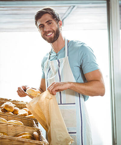 Seguro Multirriesgo de Comercio - Panadero joven sonriendo mientras se encuentra cogiendo pan en su negocio Seguro Multirriesgo de Comercio - Panadero joven sonriendo mientras se encuentra cogiendo pan en su negocio