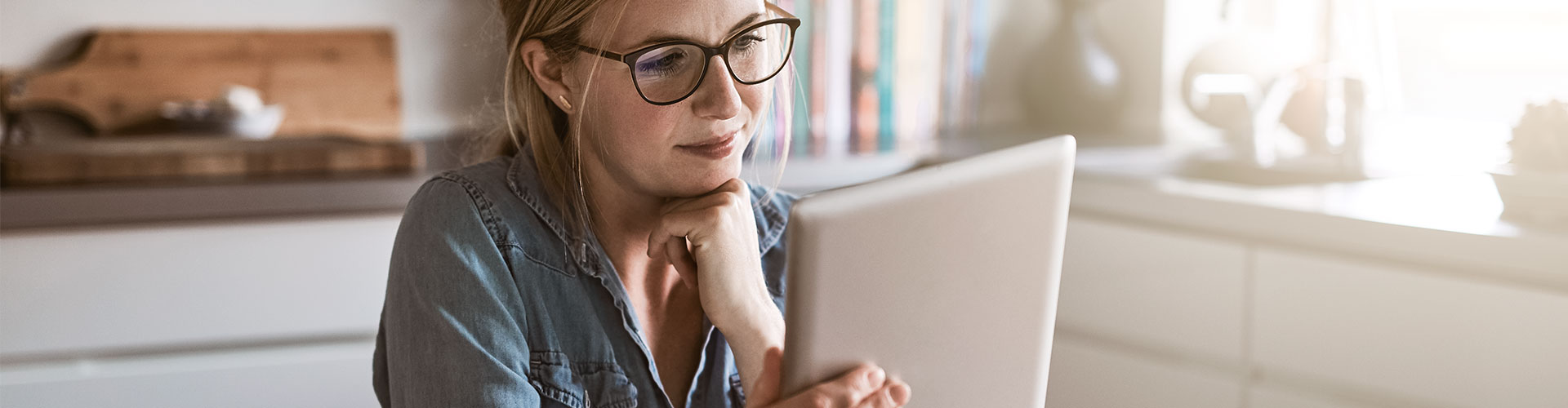 Seguro de Proteccion de Pagos - Mujer con gafas sentada en la cocina de su casa con una tablet en la mano y papeles sobre la mesa, trabajando online Seguro de Proteccion de Pagos - Mujer con gafas sentada en la cocina de su casa con una tablet en la mano y papeles sobre la mesa, trabajando online