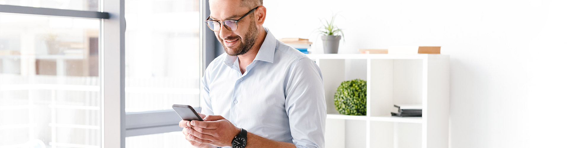 Cuenta a la Vista - Hombre de negocios con camisa y gafas sentado sobre su mesa en la oficina con un movil en la mano sonriendo