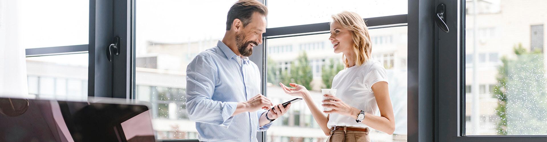 Cartera Premium - Hombre con camisa y movil en la mano, y mujer de negocios con cafe en la mano sonriendo en la oficina