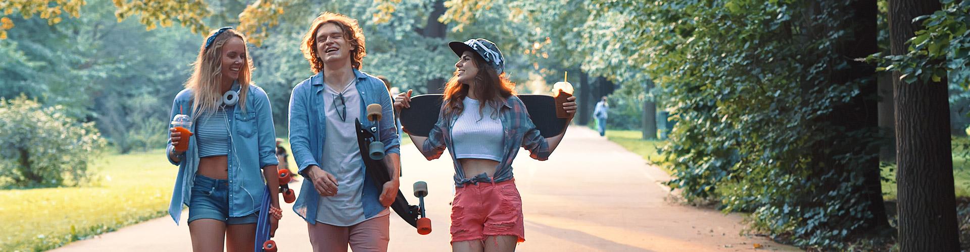 Cliente Joven In - Grupo de jovenes vestidos con ropa moderna con patinetes, sonriendo junto a un parque rodeado de arboles