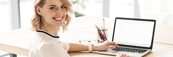 Línies ICO - Dona jove de negocis somrient mentre es troba asseguda en el seu escriptori amb el seu portàtil