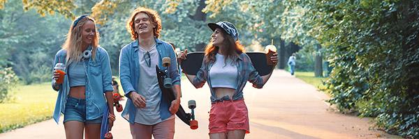 Client Joven in - Grup de joves vestits amb roba moderna amb patinets, somrient al costat d'un parc envoltat d'arbres