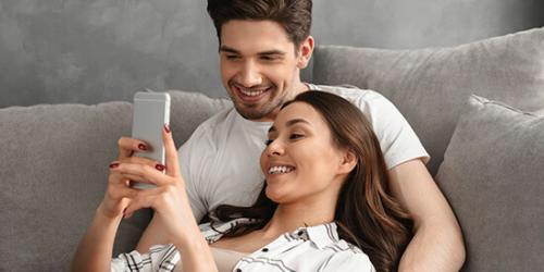 Promoció fons d'inversió - Parella jove somrient mentre estan tombats en el sofà de la seua casa mirant el mòbil i buscant fons d'inversió