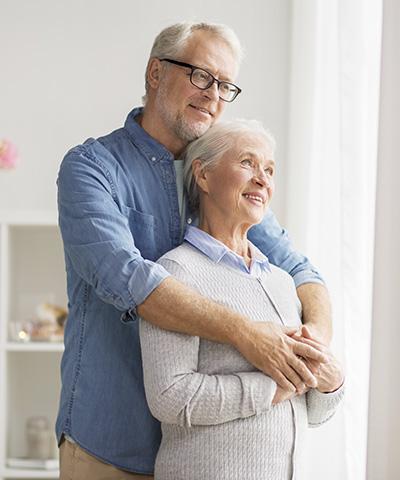 Cliente Pensionista - Una pareja senior abrazados mirando por la ventana de su casa sonriendo